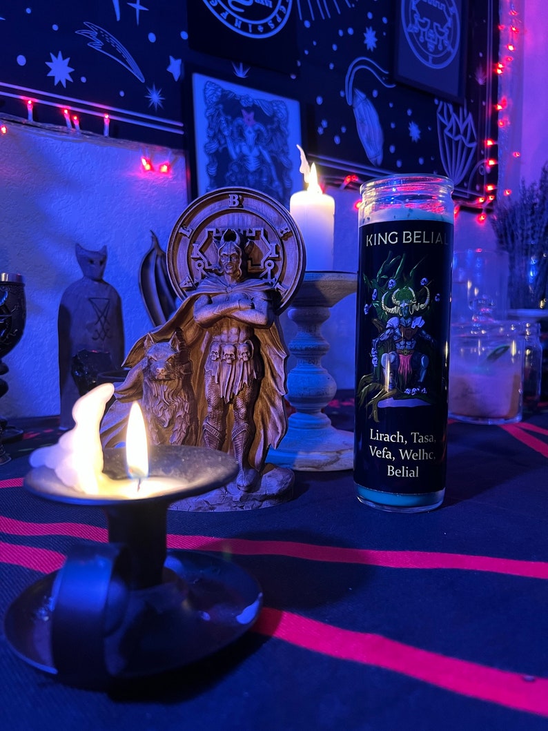 May include: A black and green candle with the text "KING BELIAL" and "Lirach, Tasa, Vefa, Welhe Belial" on the label. The candle is sitting on a table with a red and black striped tablecloth. There is a small white candle in a candlestick to the left of the candle.