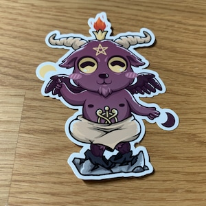 Pu&ograve; includere: Un piccolo diavolo di cartone animato viola con corna, ali e un pentagramma sulla fronte. Il diavolo indossa una tunica bianca e sta in piedi su uno scoglio. Il diavolo ha una piccola fiamma sulla testa e tiene una mezzaluna nella mano.