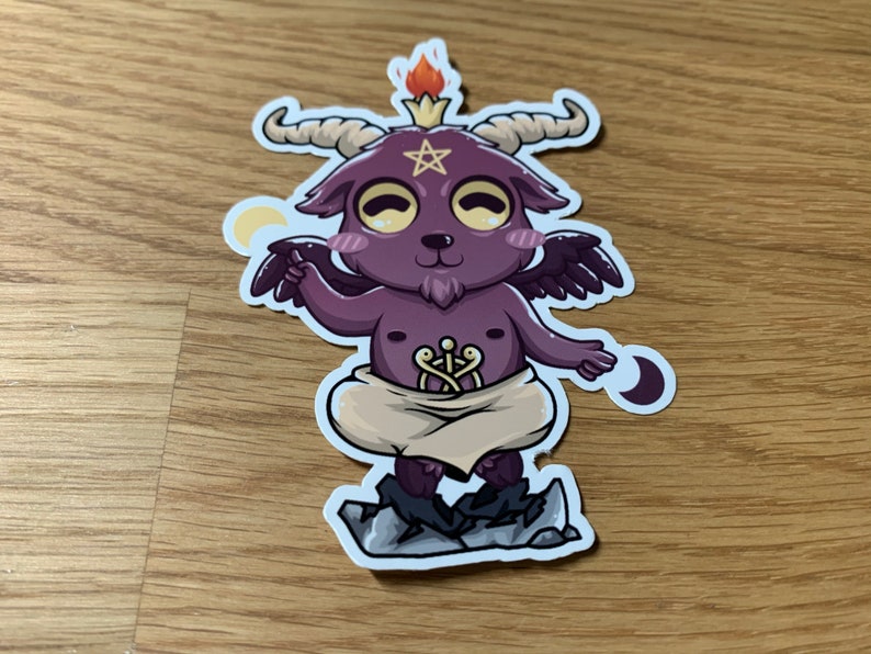 Pu&ograve; includere: Un piccolo diavolo di cartone animato viola con corna, ali e un pentagramma sulla fronte. Il diavolo indossa una tunica bianca e sta in piedi su uno scoglio. Il diavolo ha una piccola fiamma sulla testa e tiene una mezzaluna nella mano.