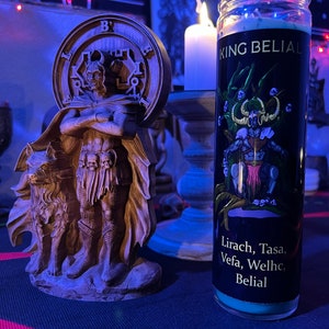 May include: A teal blue candle with a black label that reads "King Belial" and "Lirach, Tasa, Vefa, Welhc, Belial". The candle is in front of a wooden statue of a figure with a wolf.