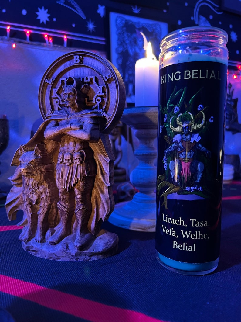 May include: A teal blue candle with a black label that reads "King Belial" and "Lirach, Tasa, Vefa, Welhc, Belial". The candle is in front of a wooden statue of a figure with a wolf.