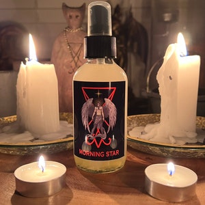 May include: A clear spray bottle with a black cap and a label that reads "MORNING STAR" with an illustration of a winged figure. The bottle is surrounded by lit candles on a wooden surface, creating a mystical ambiance.