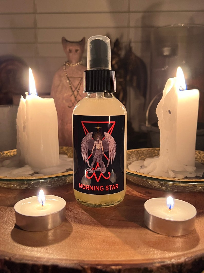 May include: A clear spray bottle with a black cap and a label that reads "MORNING STAR" with an illustration of a winged figure. The bottle is surrounded by lit candles on a wooden surface, creating a mystical ambiance.