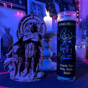 May include: A black and green candle with the text "KING BELIAL" and "Lirach, Tasa, Vefa, Welhe, Belial" on the label. The candle is in a glass jar with a black lid. The candle is sitting on a table with a dark blue and red striped tablecloth.