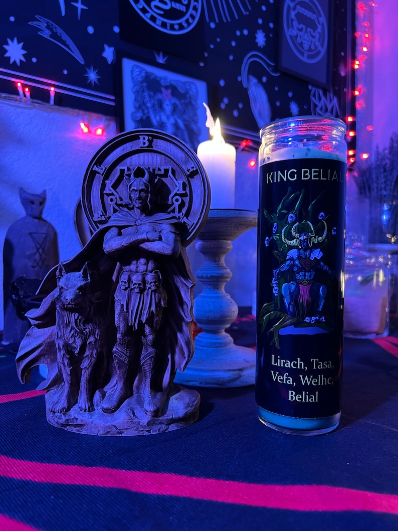 May include: A black and green candle with the text "KING BELIAL" and "Lirach, Tasa, Vefa, Welhe, Belial" on the label. The candle is in a glass jar with a black lid. The candle is sitting on a table with a dark blue and red striped tablecloth.