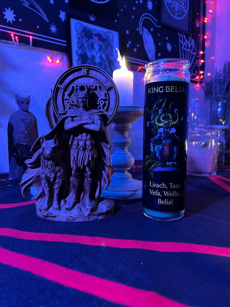 May include: A black candle with a green label that reads "King Belial" and "Lirach, Tasa, Vefa, Welhc, Belial". The candle is sitting on a black table with a red and black pattern. There is a wooden statue of a figure with a wolf to the left of the candle.