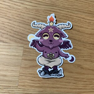 Pu&ograve; includere: Un capro di cartone animato viola con corna e ali, che indossa un panno bianco e un pentagramma sulla fronte. Il capro &egrave; in piedi su uno scoglio con una fiamma sulla testa e una mezzaluna su ogni lato.