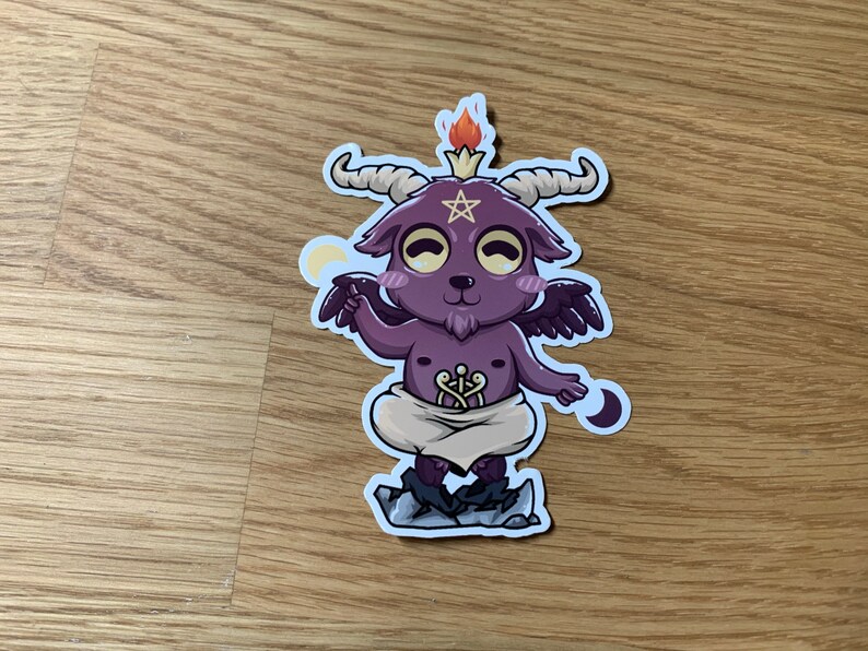 Pu&ograve; includere: Un capro di cartone animato viola con corna e ali, che indossa un panno bianco e un pentagramma sulla fronte. Il capro &egrave; in piedi su uno scoglio con una fiamma sulla testa e una mezzaluna su ogni lato.