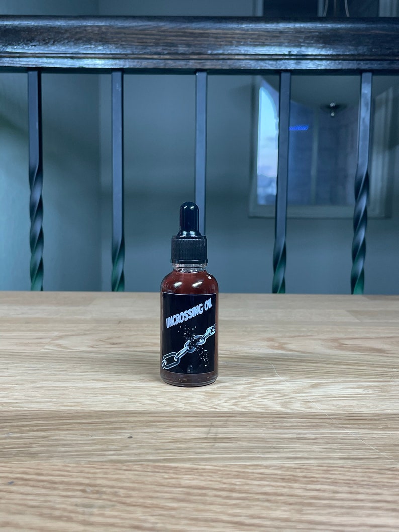 May include: A small, dark brown glass bottle with a black dropper top. The bottle has a black label with white text that reads "Uncrossing Oil" and a graphic of a chain breaking.