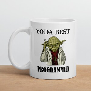 以下が含まれることがあります： スターウォーズの緑色のヨーダのキャラクターが描かれた白いセラミックマグカップ。マグカップには「ヨーダ最高のプログラマー」という黒い文字が書かれています。