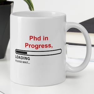 Può includere: Tazza in ceramica bianca con una grafica nera e rossa che dice "Phd in Progress, LOADING PLEASE WAIT..." con una barra di avanzamento.