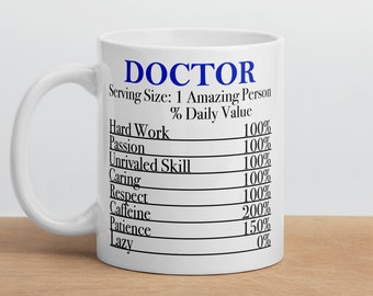 Op de afbeelding: Een witte keramische koffiemok met een zwarte en blauwe afbeelding waarop "DOCTOR" staat en de dagelijkse waarde in procenten van verschillende eigenschappen wordt vermeld, zoals hard werken, passie en cafeïne.