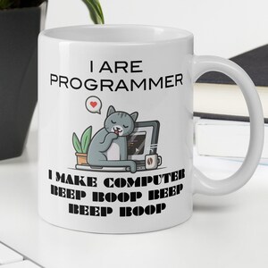 May include: White ceramic mug with a black handle. The mug has the text "I ARE PROGRAMMER" above a cartoon cat sleeping in front of a computer. Below the cat is the text "I MAKE COMPUTER BEEP BOOP BEEP BEEP BOOP". A small heart icon is above the cat.