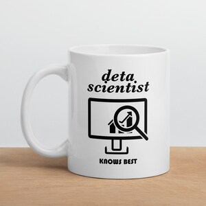 May include: White ceramic mug with the words "data scientist" and a graphic of a computer monitor with a magnifying glass over a graph. The text "KNOWS BEST" is below the monitor graphic. The mug is on a wooden surface.