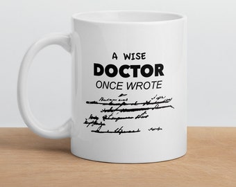 Op de afbeelding: Witte keramische mok met een zwarte inscriptie die luidt "Een wijze dokter schreef ooit" met een handgeschreven recept eronder.