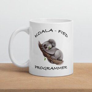 May include: White ceramic mug featuring a koala bear illustration on a tree branch, with the text "KOALA-FIED PROGRAMMER" in black. The koala is gray and white, holding a green leaf. The mug has a curved handle and is on a wooden surface.