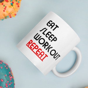 May include: White ceramic mug with black text that reads "Eat Sleep Workout Repeat" with the word "Repeat" in red. The mug is surrounded by colourful doughnuts.