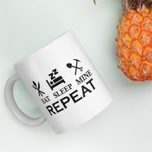 Peut inclure: Une tasse en céramique blanche avec un motif noir qui dit "Manger Dormir Mine Répéter" avec des icônes d'une fourchette et d'un couteau, d'un lit et d'une pioche.