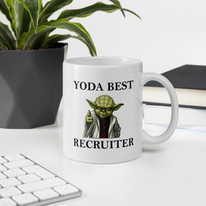Pu&ograve; includere: Una tazza in ceramica bianca con un'immagine in bianco e verde di Yoda di Star Wars. Il testo "YODA BEST RECRUITER" &egrave; stampato sulla tazza.