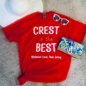 Pode incluir: Camiseta vermelha com texto branco que diz "CREST is the BEST Wildwood Crest, New Jersey". Um chap&eacute;u de sol branco e &oacute;culos de sol est&atilde;o sobre a camiseta. Uma pequena bolsa com estampa tropical est&aacute; do lado direito da camiseta.