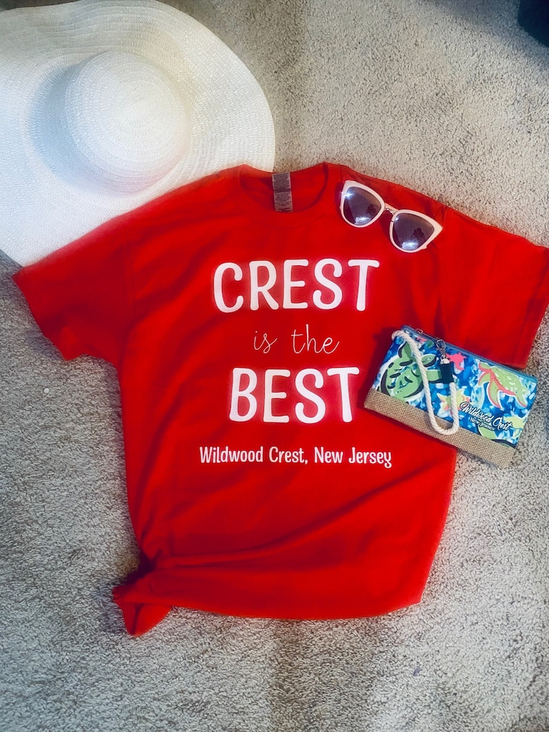 Pode incluir: Camiseta vermelha com texto branco que diz "CREST is the BEST Wildwood Crest, New Jersey". Um chap&eacute;u de sol branco e &oacute;culos de sol est&atilde;o sobre a camiseta. Uma pequena bolsa com estampa tropical est&aacute; do lado direito da camiseta.