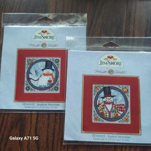 May include: Two Jim Shore by Mill Hill cross stitch kits in clear plastic packaging. The left kit is JS14-9102 Southern Snowman, and the right is JS14-9103 Eastern Snowman. Each kit features a snowman design with a 5 inch by 5 inch finished size.
