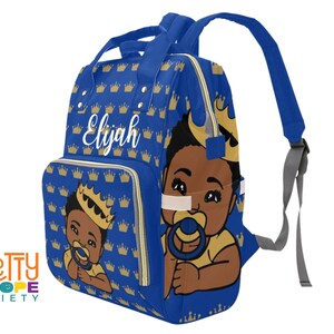 Puede incluir: Bolso para pa&ntilde;ales azul real con un patr&oacute;n de coronas doradas. El bolso presenta una ilustraci&oacute;n de un beb&eacute; con corona y chupete. El nombre "Elijah" est&aacute; escrito en blanco. Tiene un asa superior y correas ajustables.