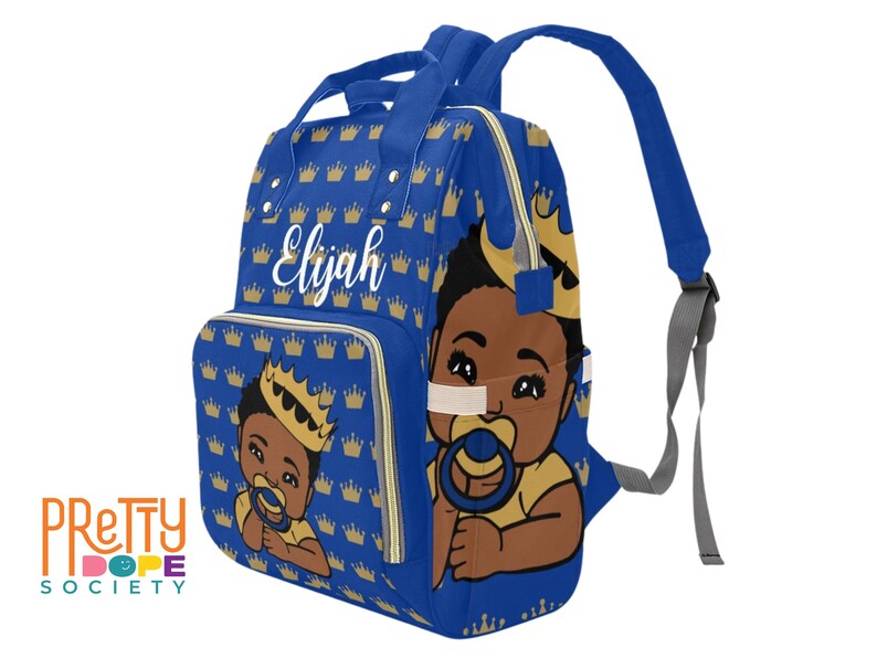 Puede incluir: Bolso para pa&ntilde;ales azul real con un patr&oacute;n de coronas doradas. El bolso presenta una ilustraci&oacute;n de un beb&eacute; con corona y chupete. El nombre "Elijah" est&aacute; escrito en blanco. Tiene un asa superior y correas ajustables.