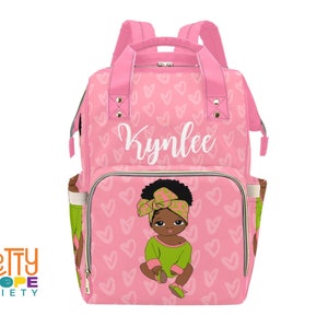 May include: Pink baby backpack with a heart pattern and the name "Kynlee" in white script. The front pocket features a cartoon illustration of a baby girl in green and brown. The bag has pink straps and handles.