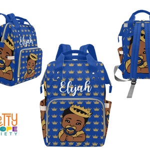 Puede incluir: Bolso de pa&ntilde;ales azul con un patr&oacute;n de coronas doradas repetidas. El bolso presenta una ilustraci&oacute;n de un beb&eacute; con una corona y un chupete. El nombre "Elijah" est&aacute; escrito en blanco en la parte delantera.