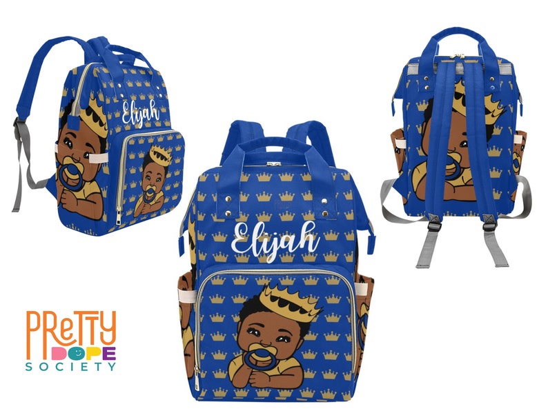 Puede incluir: Bolso de pa&ntilde;ales azul con un patr&oacute;n de coronas doradas repetidas. El bolso presenta una ilustraci&oacute;n de un beb&eacute; con una corona y un chupete. El nombre "Elijah" est&aacute; escrito en blanco en la parte delantera.