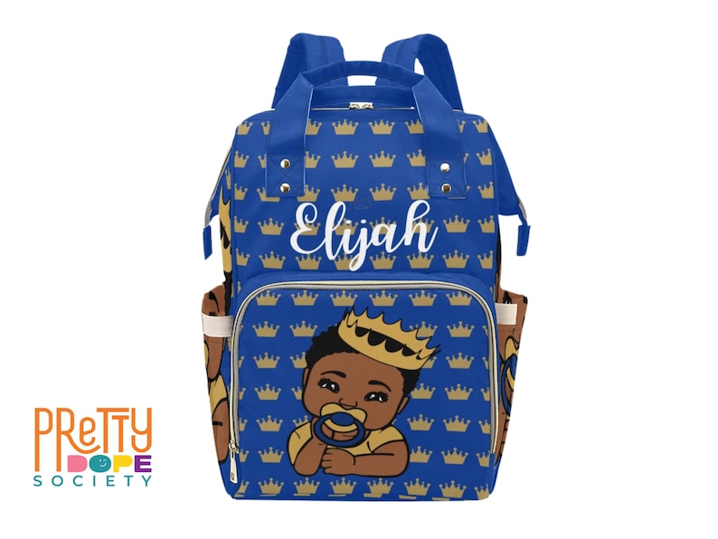 Puede incluir: Un bolso para pa&ntilde;ales azul real con un estampado de corona dorada y el nombre "Elijah" en escritura blanca. El bolso presenta una ilustraci&oacute;n de dibujos animados de un beb&eacute; pr&iacute;ncipe con una corona y un chupete. El bolso tiene m&uacute;ltiples bolsillos y correas.