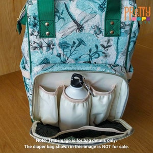 Puede incluir: Un bolso de pa&ntilde;ales turquesa y blanco con un estampado de lib&eacute;lulas y flores. El bolso tiene correas verdes y un bolsillo frontal con una botella de agua blanca. El bolso tiene m&uacute;ltiples bolsillos para guardar cosas. El texto en el bolso dice "PRETTY SOCIETY".