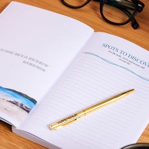 May include: An open notebook with lined pages and the text "SPOTS TO DISCOVER" in blue. A gold pen rests on the page. The notebook also features a quote by Ralph Waldo Emerson. Black glasses are in the background.