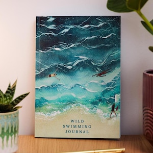 May include: A journal with a watercolor illustration of swimmers in the ocean. The cover features a beach scene with figures in the water and the text "Wild Swimming Journal". The color palette includes shades of blue, green, and white.