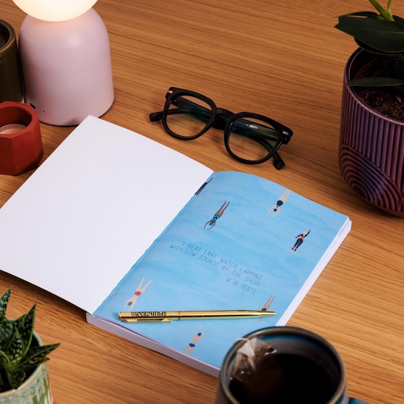 May include: An open notebook with a blue illustration of swimmers in a lake, a gold pen, and black eyeglasses on a wooden surface. A quote by W.B. Yeats is printed on the page. A pink lamp and a cup of tea are also visible.
