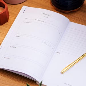 May include: Open swim log journal with the title "SWIM LOG" at the top. The journal has sections for date, water conditions, weather, mood, and a sketch area. A gold pen rests on the right page.