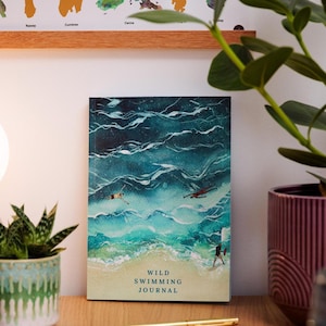 May include: A journal with a watercolor illustration of ocean waves and swimmers. The cover reads "Wild Swimming Journal" in a serif font. The journal is on a wooden surface with a plant in a decorative pot.