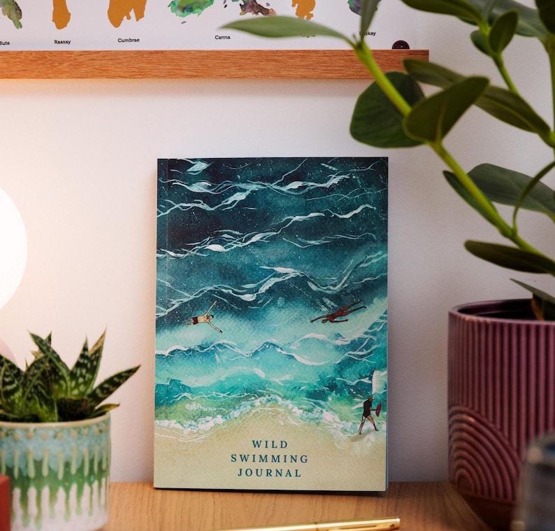 May include: A journal with a watercolor illustration of ocean waves and swimmers. The cover reads "Wild Swimming Journal" in a serif font. The journal is on a wooden surface with a plant in a decorative pot.