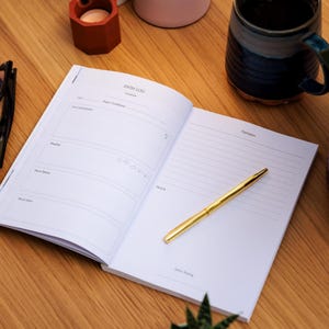 May include: An open notebook with the words "SWIM LOG" at the top, a gold pen, and a mug on a wooden surface. The notebook has sections for recording details like date, weather, and mood. The pen is centered on the right page.