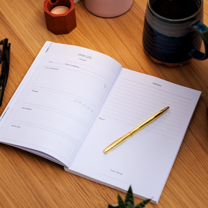 May include: An open notebook with the words "SWIM LOG" at the top, a gold pen, and a mug on a wooden surface. The notebook has sections for recording details like date, weather, and mood. The pen is centered on the right page.
