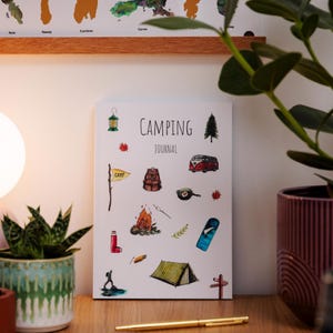 May include: A camping journal with a white cover featuring camping-themed illustrations. The journal includes images of a tent, backpack, and campfire. The words "CAMPING JOURNAL" are printed in black. A gold pen is on the wooden surface.