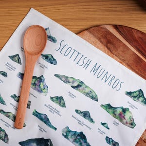 May include: A wooden spoon rests on a white tea towel featuring a map of Scottish Munros. The towel displays watercolor illustrations of mountains with their names and elevations in meters. The spoon is light brown and the towel is on a wooden cutting board.