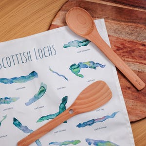 May include: A white tea towel with a watercolor map of Scottish lochs, with two wooden spoons and a wooden cutting board. The tea towel has the words "Scottish Lochs" in blue. The spoons are light brown.