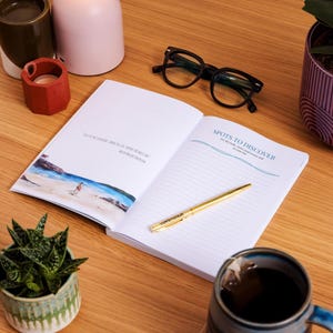 May include: An open journal with the text "Spots to Discover" and a gold pen on a wooden surface. The journal has lined pages and a watercolor illustration. Black eyeglasses and a mug of tea are also visible.