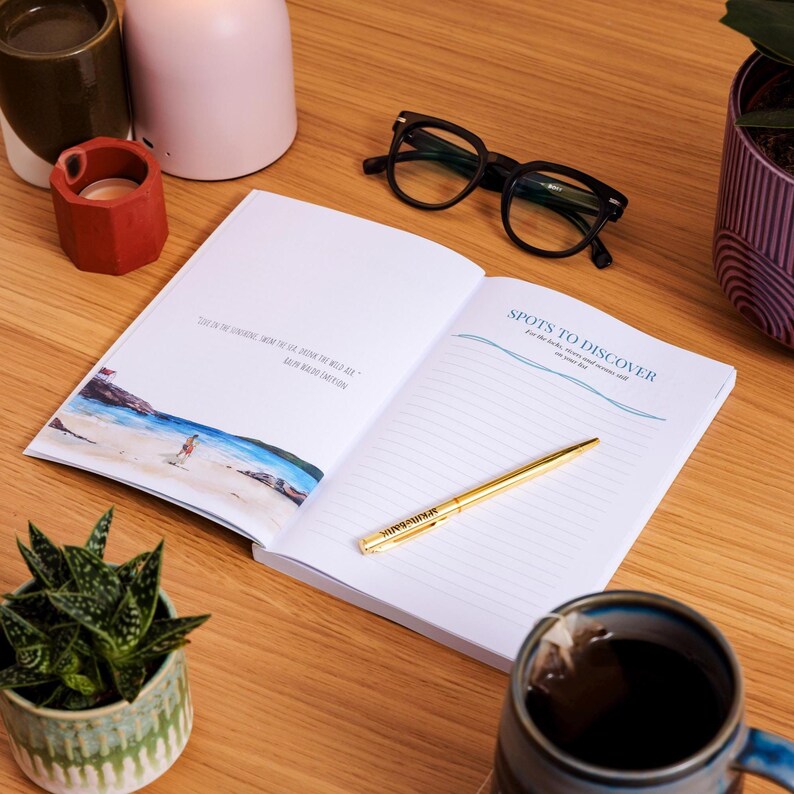 May include: An open journal with the text "Spots to Discover" and a gold pen on a wooden surface. The journal has lined pages and a watercolor illustration. Black eyeglasses and a mug of tea are also visible.