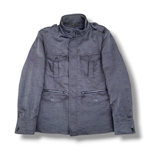 K&ouml;nnte beinhalten: Eine dunkelgraue Utility-Jacke mit hohem Kragen und Rei&szlig;verschluss-Detail, Schulterklappen und langen &Auml;rmeln mit gekn&ouml;pften Manschetten. Die Vorderseite hat zwei Brusttaschen mit Knopfklappen und zwei untere Rei&szlig;verschlusstaschen mit Knopfklappen.