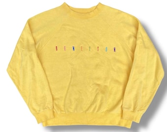 Puede incluir: Una sudadera amarilla con la palabra "BENETTON" bordada en colores arcoíris. La parte superior de manga larga tiene cuello redondo y un ajuste relajado. La prenda está hecha de un material suave.