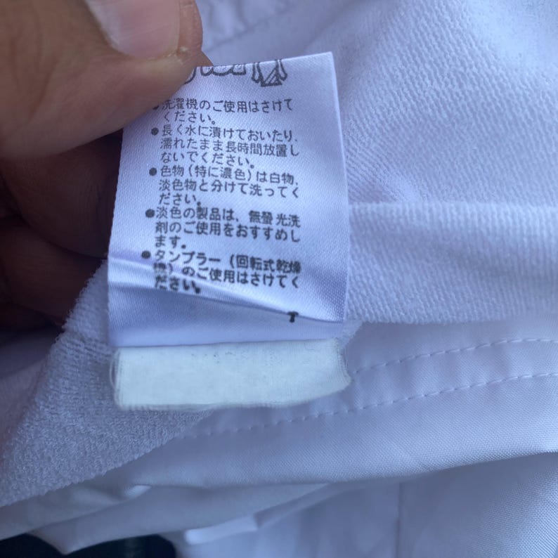 May include: A close-up of a white fabric with a sewn seam and a care label attached. The label has Japanese text with care instructions, including washing and drying advice.