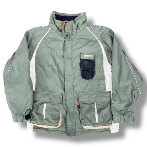 Könnte beinhalten: Salbeigrüner Jacke mit weißen und beigen Akzenten. Die Jacke hat einen hohen Kragen, mehrere Taschen und eine schwarze Netztasche auf der Brust. Sie hat einen Reißverschluss und orangefarbene Reißverschlussanhänger. Größe L.